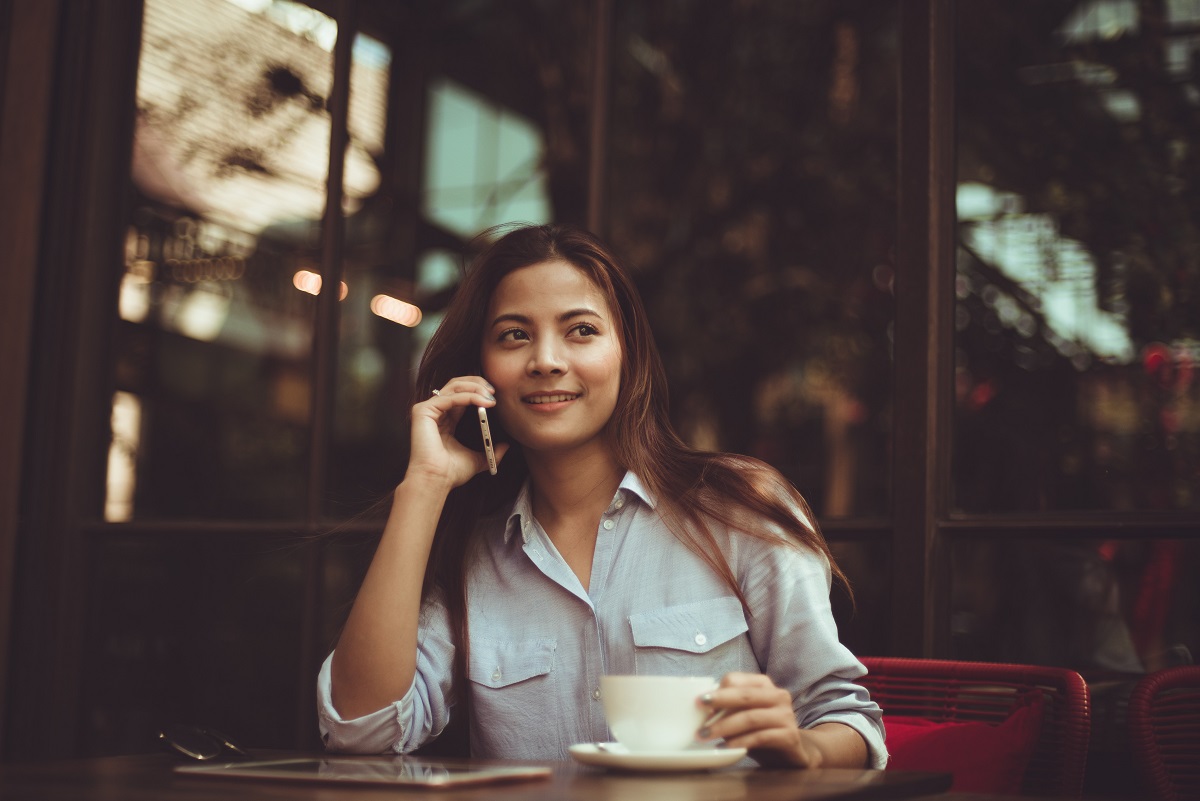 young female Asian American professional sitting at cafe on cellphone drinking coffee
